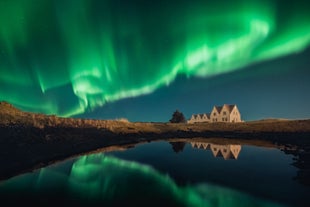 A traditional Icelandic house reflects in still water beneath the vibrant green northern lights in a clear night sky.