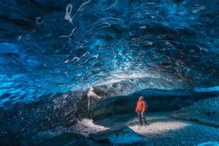 A hiker in a bright winter jacket standing in the middle of a glowing blue ice tunnel.