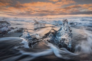 Pieces of ice from towering icebergs wash ashore on Diamond Beach, with waves flowing around them on the black sand.