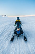A person rides a snowmobile across a snowy glacier in Iceland under a clear blue sky during a winter adventure.