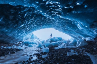 A person stands inside a Katla blue ice cave in Iceland, surrounded by glistening ice walls and frozen boulders.