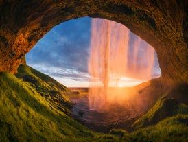 Behind the falls of Seljalandsfoss in Iceland, sunset turns the water golden against green mossy cliffs.