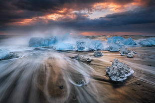 Waves rush around crystal-clear ice chunks on Diamond Beach at sunrise, with clouds and warm light glowing on the horizon.