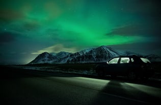 In Iceland, a car is parked under the northern lights, with snowy mountains in the background.