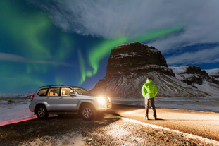 A traveler watches the northern lights dance above Mount Lomagnupur in Iceland beside a car on a snowy roadside.