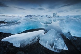 Icebergs floating in Jokulsarlon Glacier Lagoon with reflections on still water and snow-covered mountains in the distance.